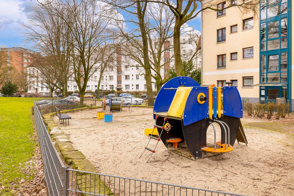 Spielplatz mit Klettergerüst und Bänken neben Wohngebäuden und geparkten Autos.