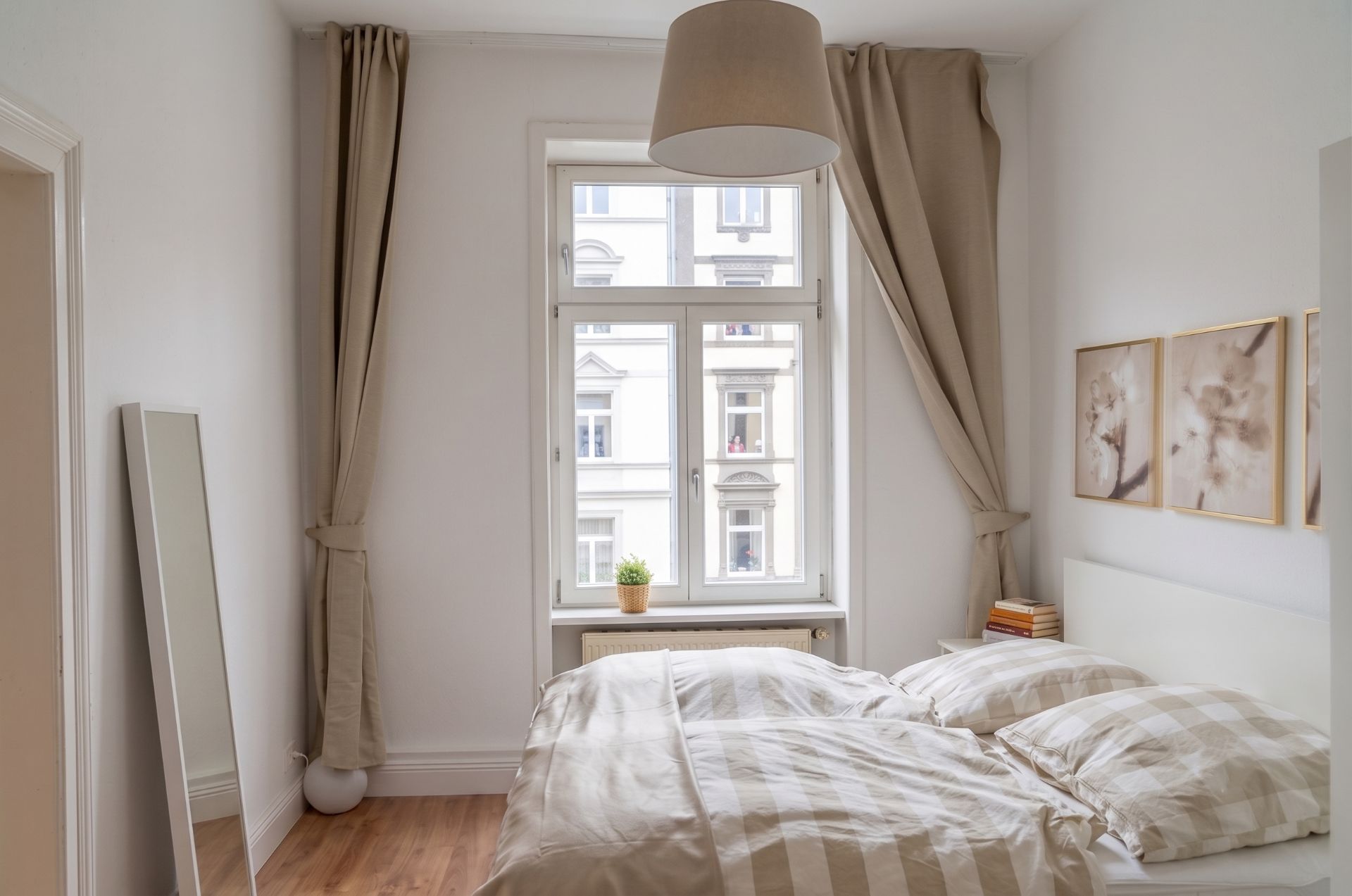Helles Schlafzimmer mit Doppelbett, großem Fenster, beigen Vorhängen und Wandbildern.
