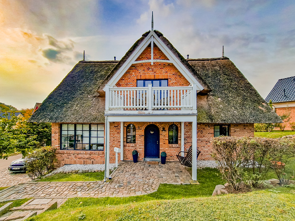 Backsteinhaus mit Reetdach, Balkon und rundem blauen Eingangstor, umgeben von Garten.