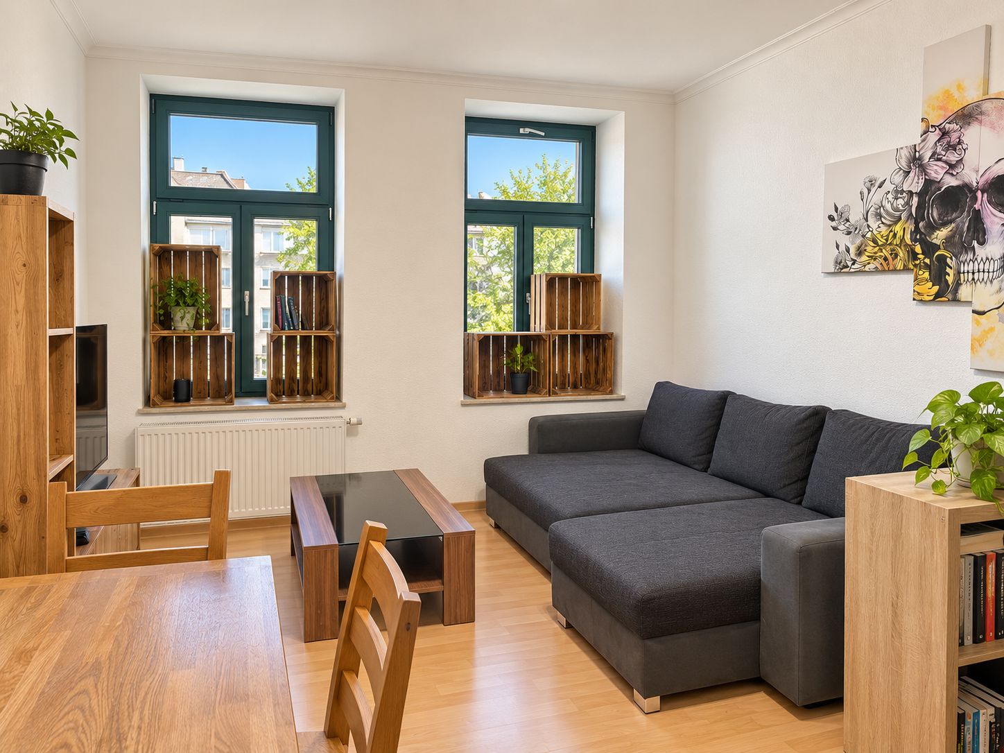 Helles Wohnzimmer mit grauem Sofa, Holzmöbeln, Pflanzen und zwei Fenstern.