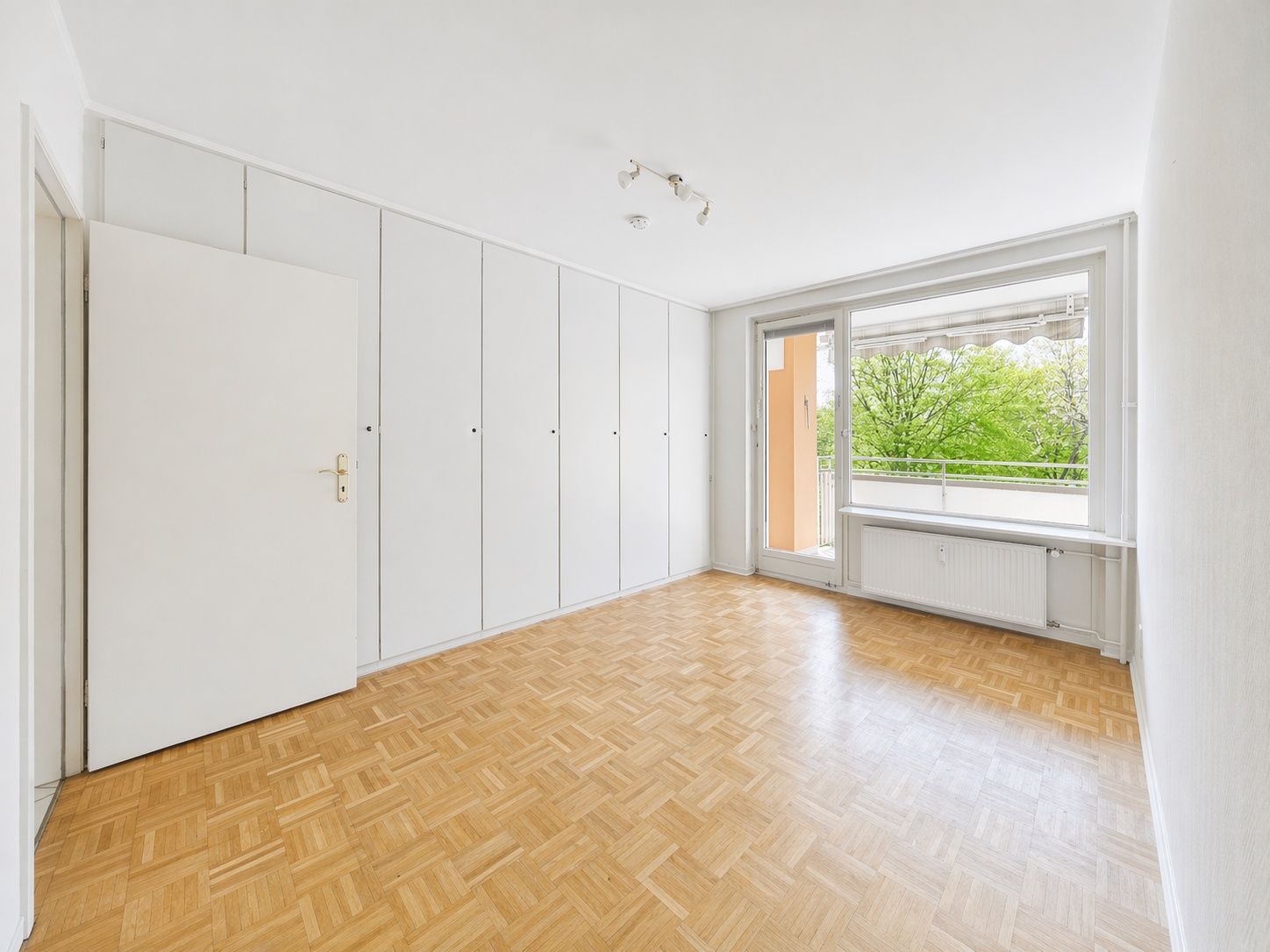 Heller, leerer Raum mit Parkettboden, weißen Einbauschränken und großer Fensterfront mit Balkonzugang.