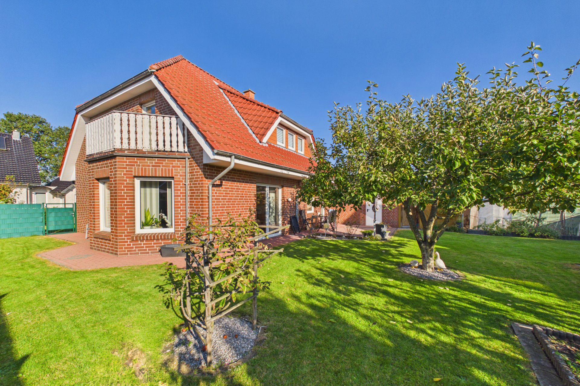 Backstein-Einfamilienhaus mit rotem Dach, Balkon und Garten mit Bäumen.