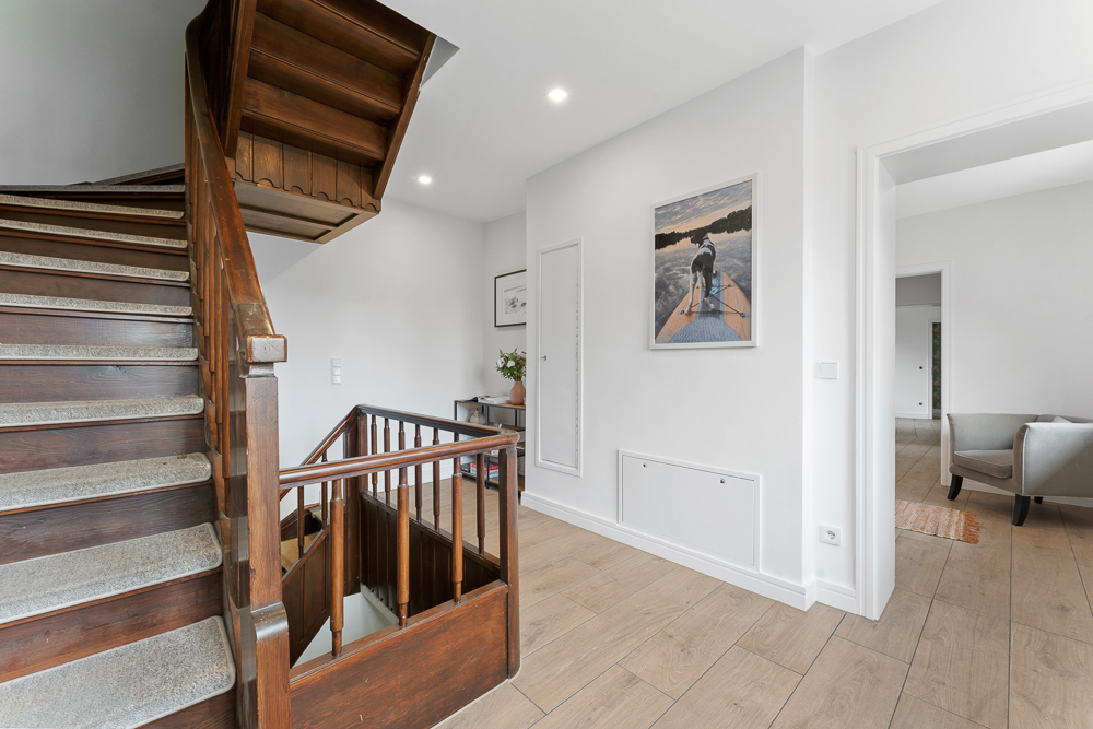 Flur mit hölzerner Treppe, hellem Boden und Wandbild, rechts daneben Zugang zum Wohnzimmer.