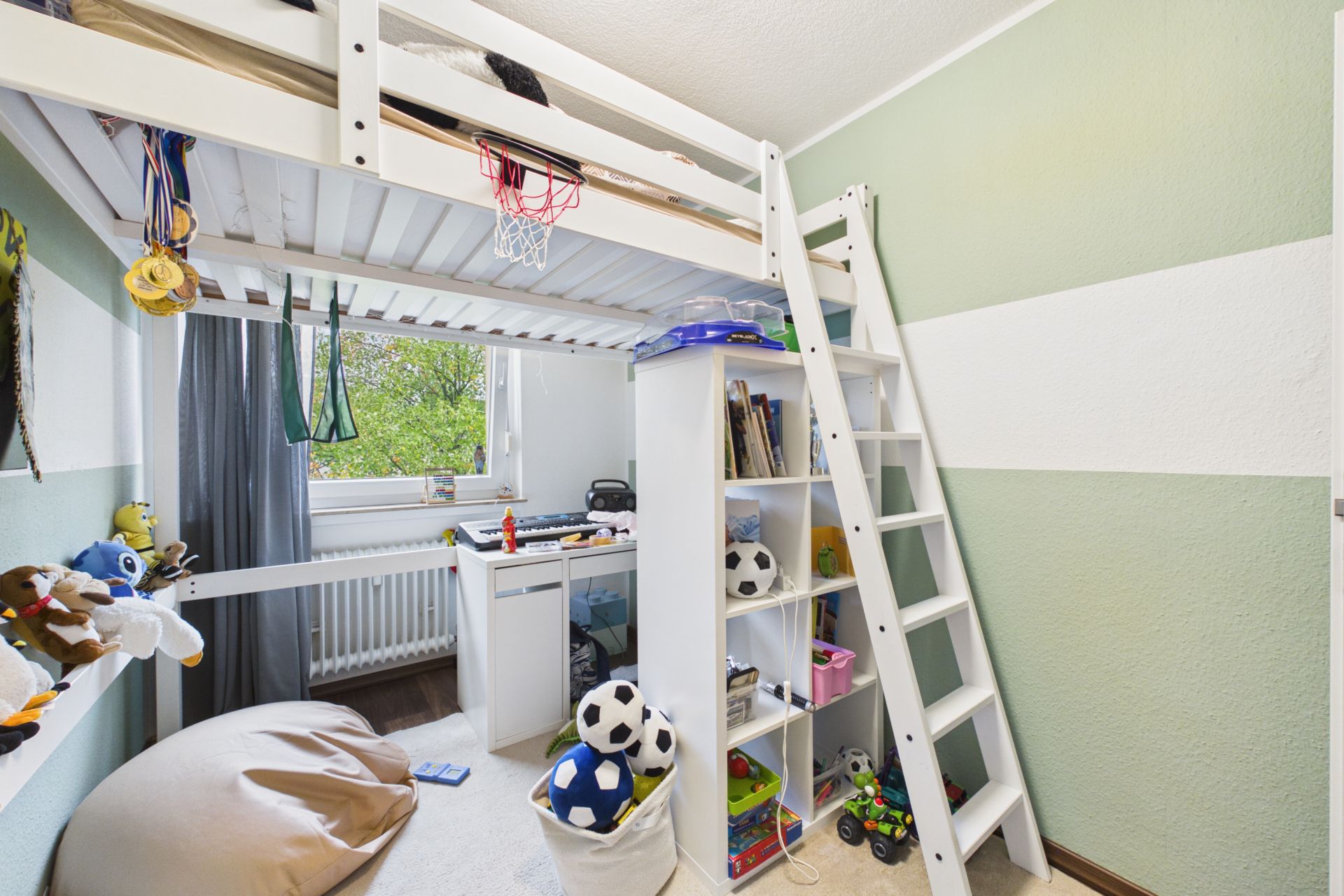 Kinderzimmer mit Hochbett, Regal, Spielzeug und Schreibtisch vor dem Fenster.