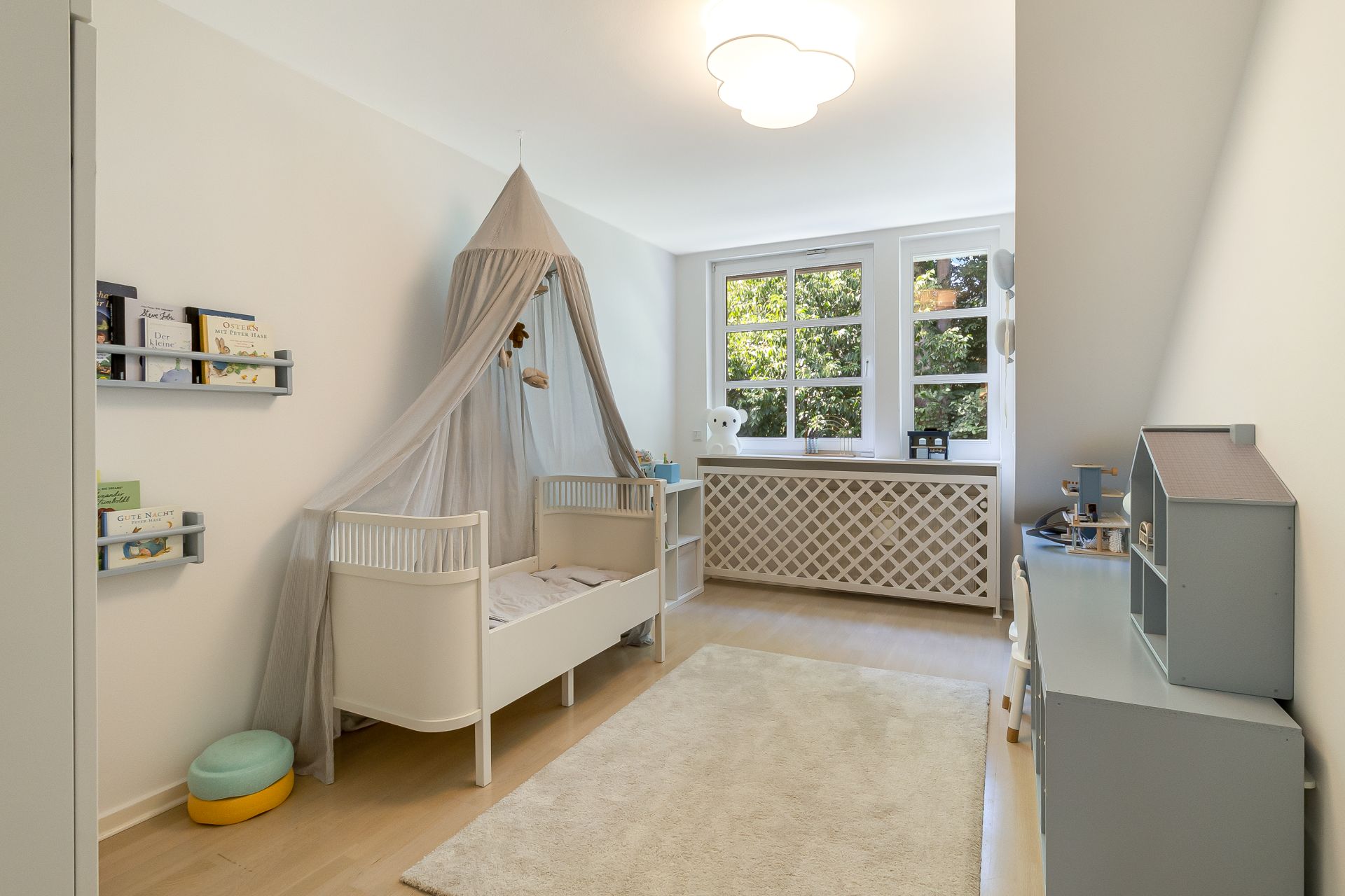Helles Kinderzimmer mit Babybett, Bücherregalen und großem Fenster, hell eingerichtet.