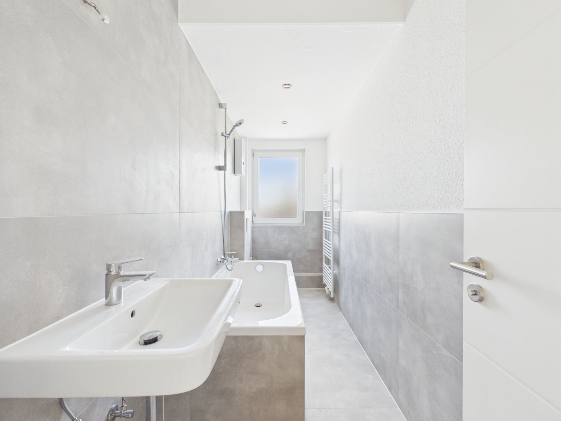 Helles Badezimmer mit Waschbecken, Badewanne, grauen Fliesen und Fenster am Ende des Raums.