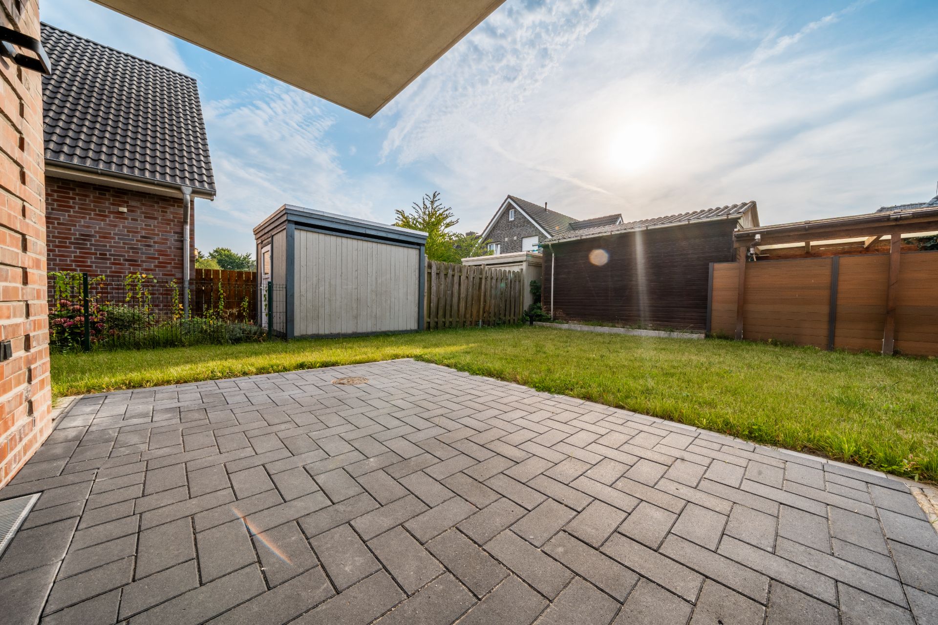 Gepflasterte Terrasse mit angrenzendem Rasen, Schuppen und Holzzaun im Garten.