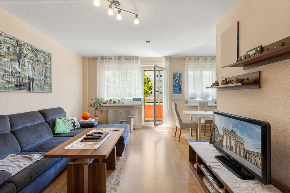 Helles Wohnzimmer mit Sofa, Couchtisch, Fernseher, Esstisch und Zugang zum Balkon.