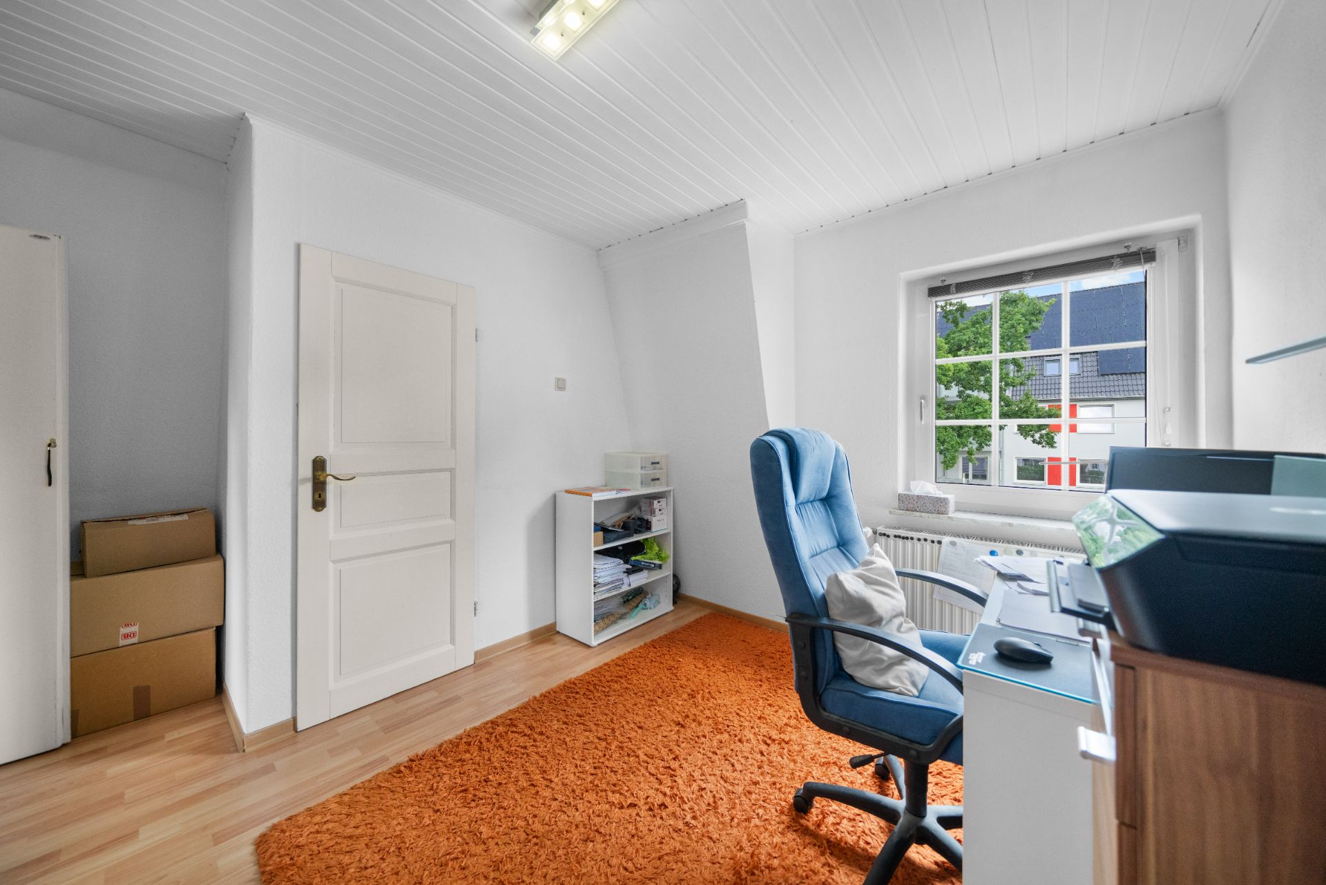 Helles Arbeitszimmer mit Schreibtisch, Bürostuhl, Regal und orangefarbenem Teppich vor einem Fenster.