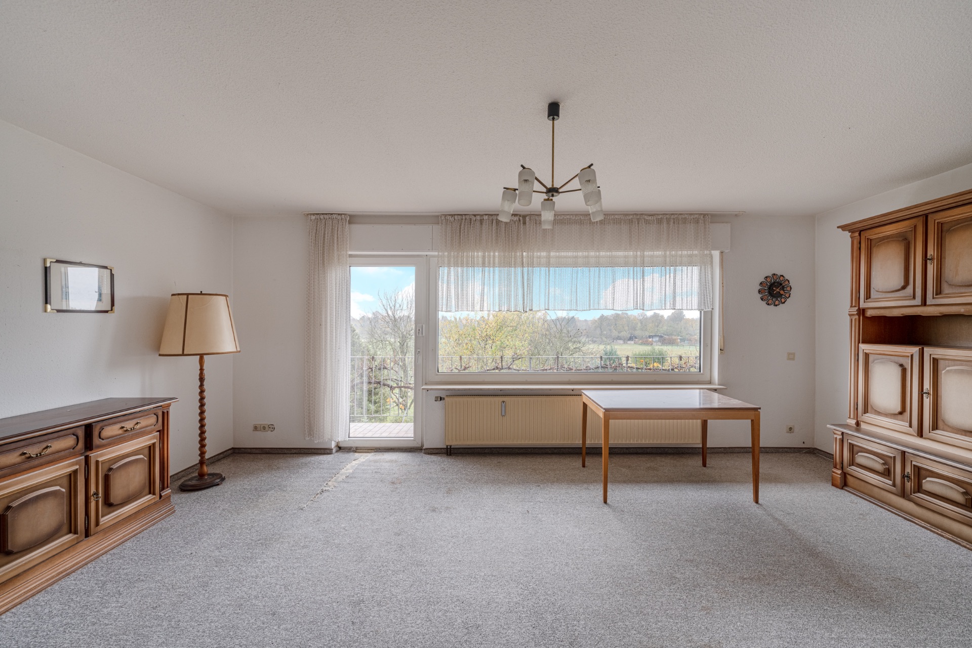 Wohnzimmer mit Teppichboden, Holzmöbeln, Stehlampe und großem Fenster mit Blick nach draußen.