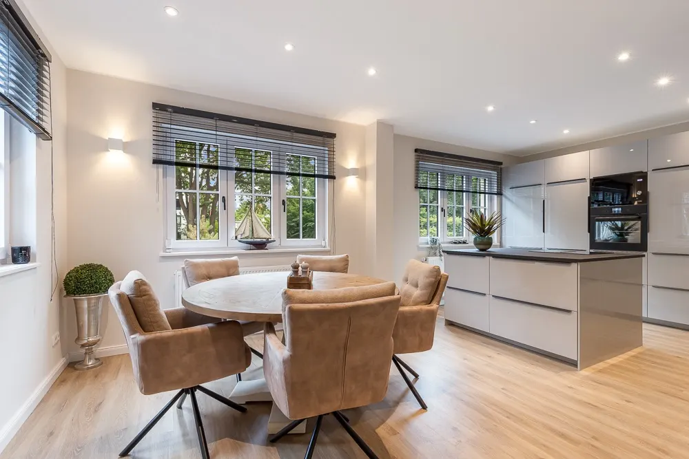 Moderne Küche mit Essbereich, runder Tisch und sechs beige Stühle, große Fenster und Holzboden.