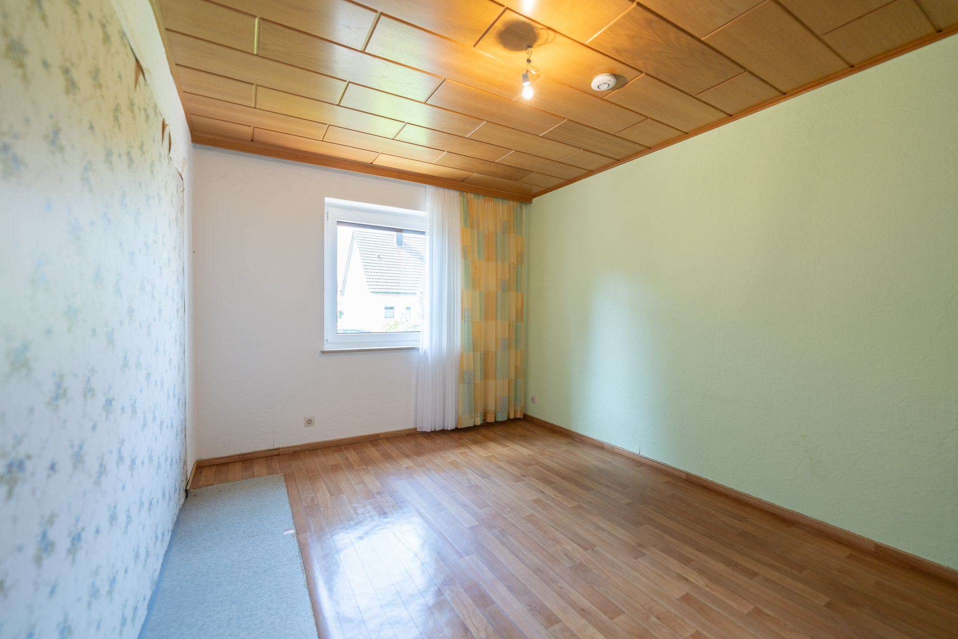 Leeres Zimmer mit Holzdecke, Parkettboden, hellem Fenster und gemusterter Tapete.