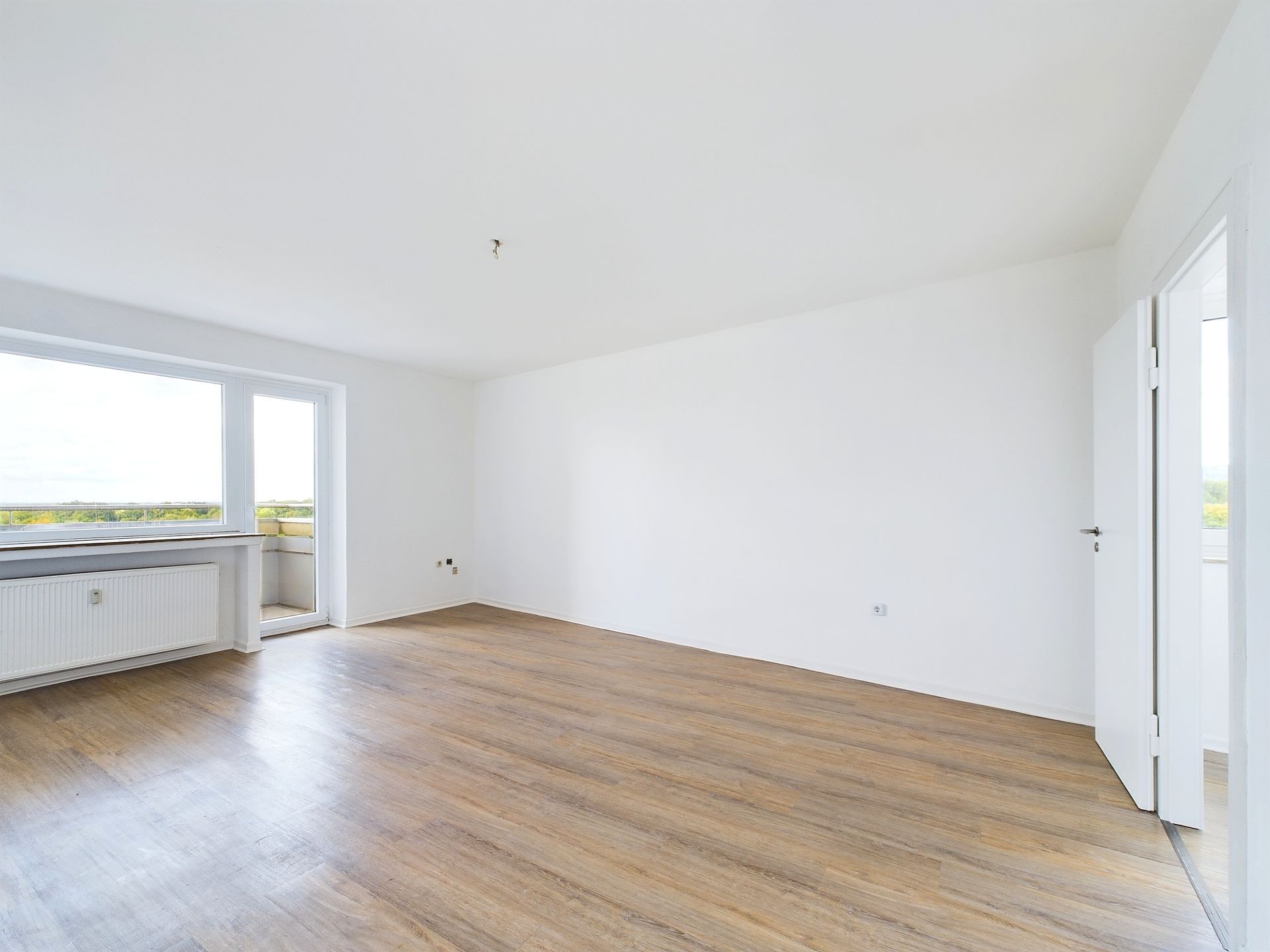 Helles, leeres Zimmer mit Holzfußboden, weißen Wänden und großem Fenster.