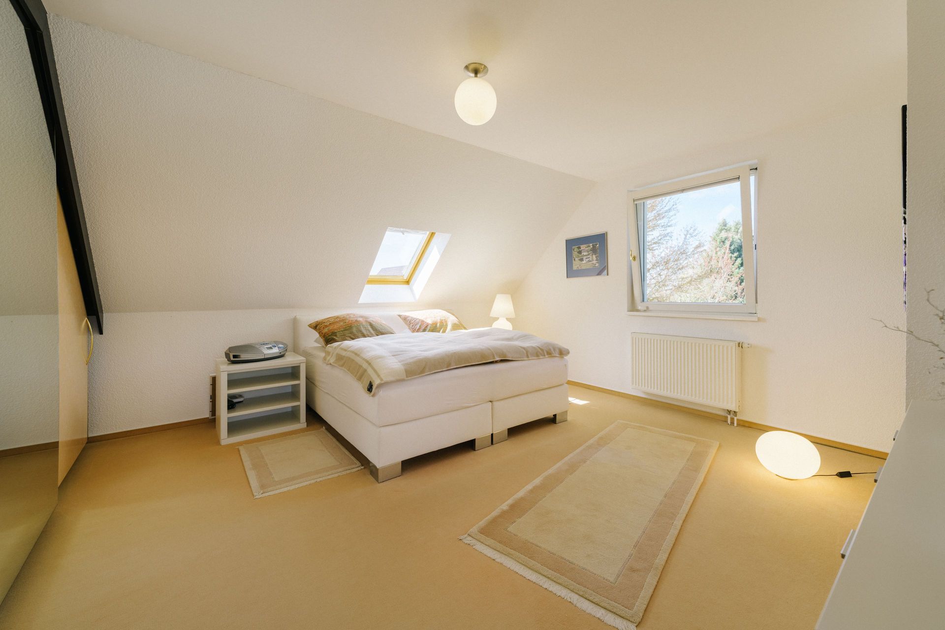 Helles Schlafzimmer mit Doppelbett, Teppichen, Nachttisch, Lampe und schrägem Fenster.