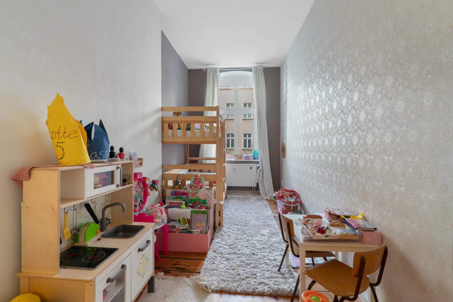 Kinderzimmer mit Hochbett, Spielküche, Spielzeug und Schreibtisch, großes Fenster im Hintergrund.