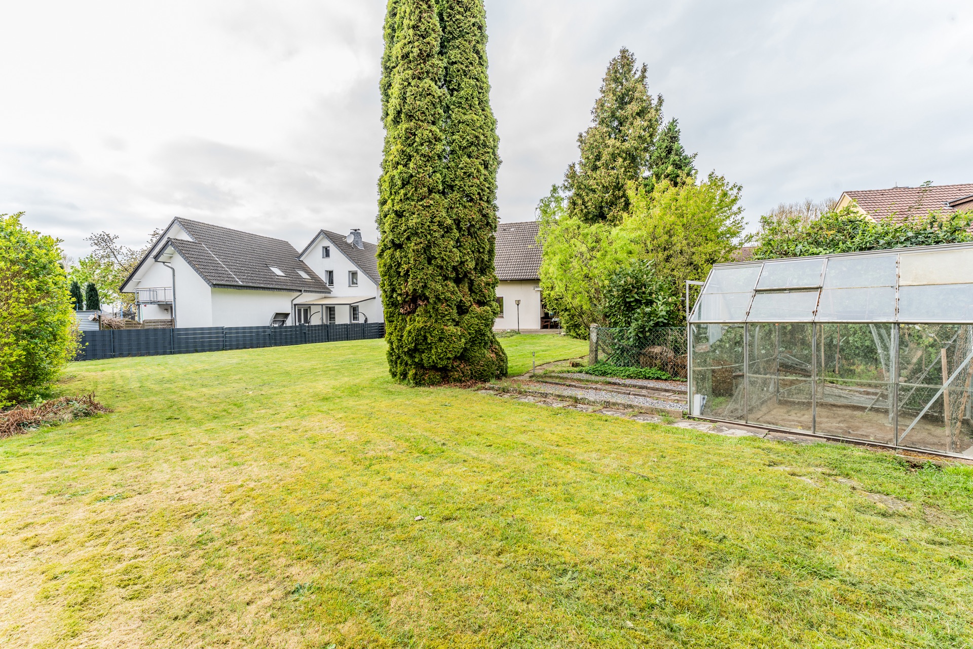 Großer Garten mit Rasen, hohen Bäumen und einem Gewächshaus neben Wohnhäusern.