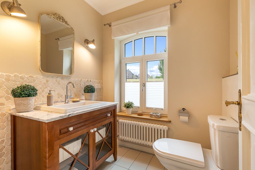 Helles Badezimmer mit Fenster, Waschbecken, Spiegel, WC und Pflanzen auf der Ablage.