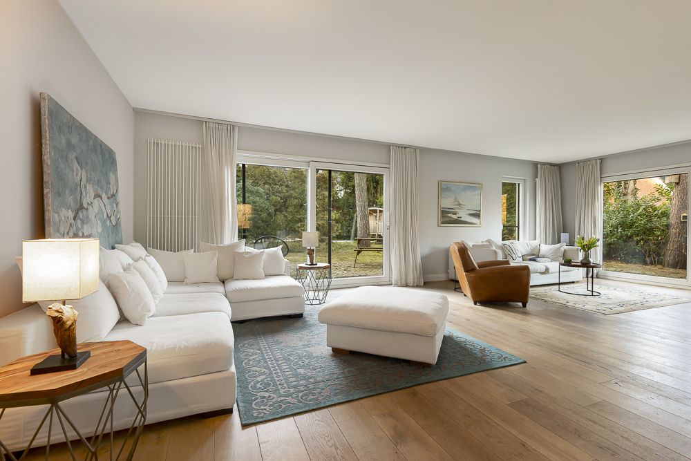 Helles Wohnzimmer mit Sofa, Sessel, Teppich und großen Fenstern mit Blick ins Grüne.