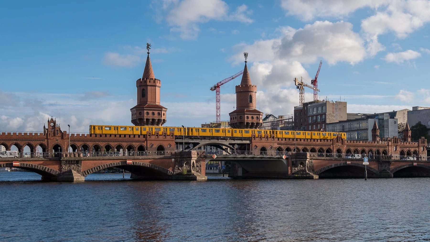 Backsteinbrücke mit zwei Türmen, gelber Zug fährt darüber, Wasser im Vordergrund.