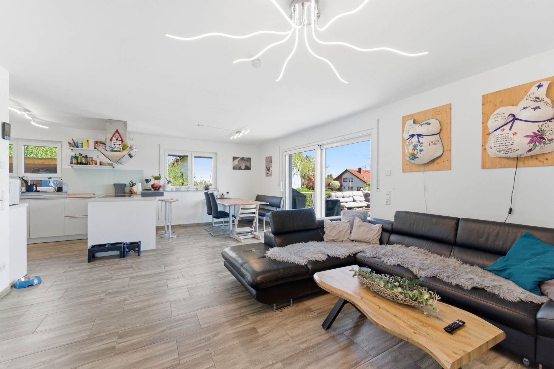 Helles Wohnzimmer mit schwarzem Ecksofa, Holztisch, offener Küche und Essbereich mit großen Fenstern.