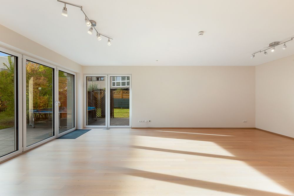 Helles, leeres Wohnzimmer mit Holzboden und großen Glas-Schiebetüren zum Garten.