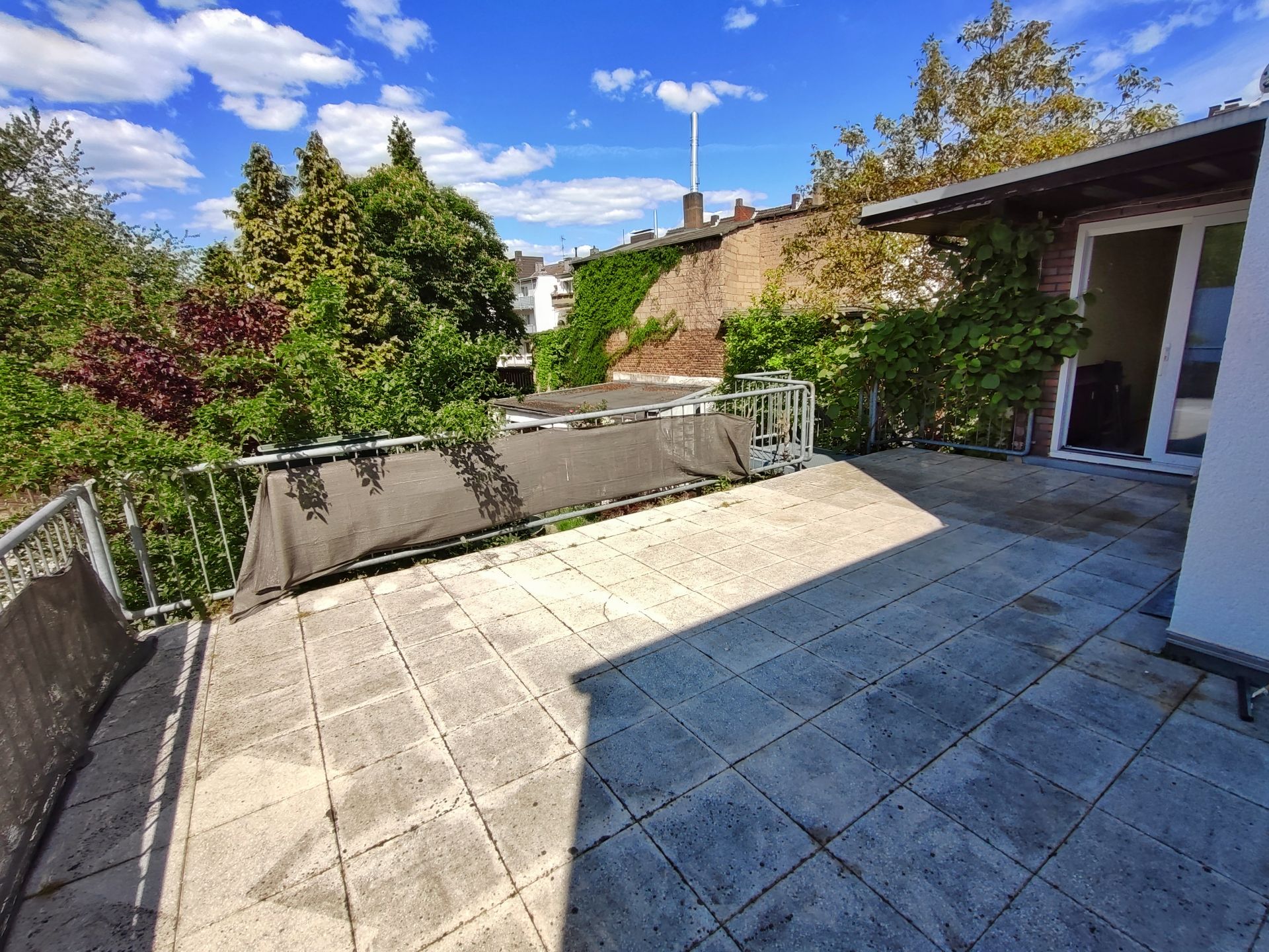 Große Terrasse mit Fliesenboden, Geländer und Blick auf grüne Bäume, angrenzend an ein Wohnhaus.