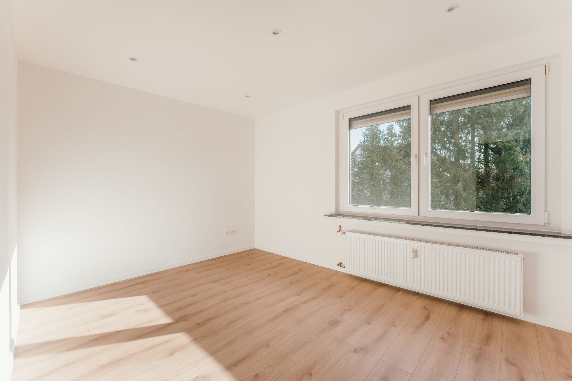Leeres Zimmer mit hellem Holzfußboden, großem Fenster und Heizkörper unter dem Fenster.