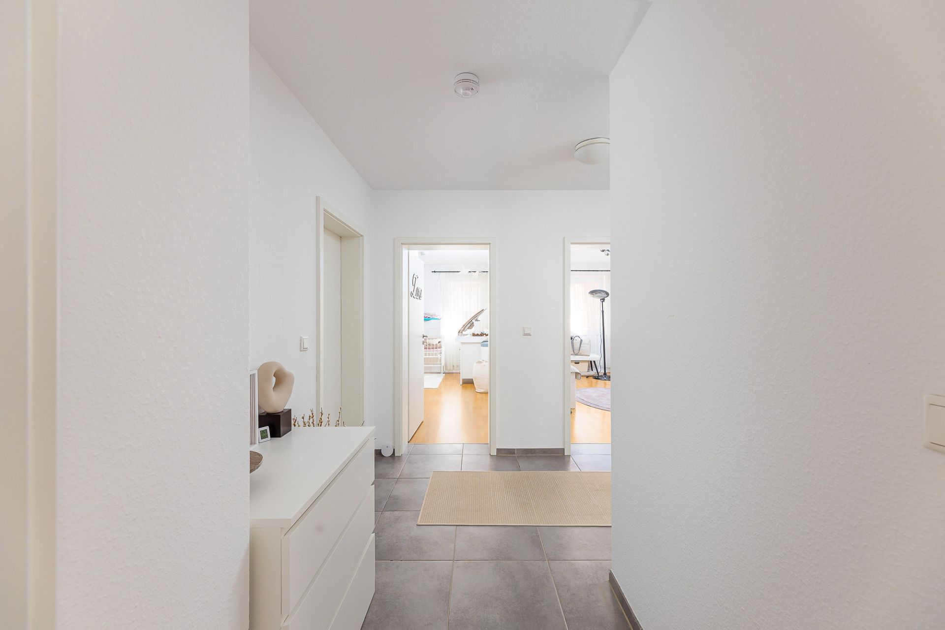 Heller Flur mit Fliesenboden, Kommode links und Blick in zwei helle Zimmer mit Holzboden.