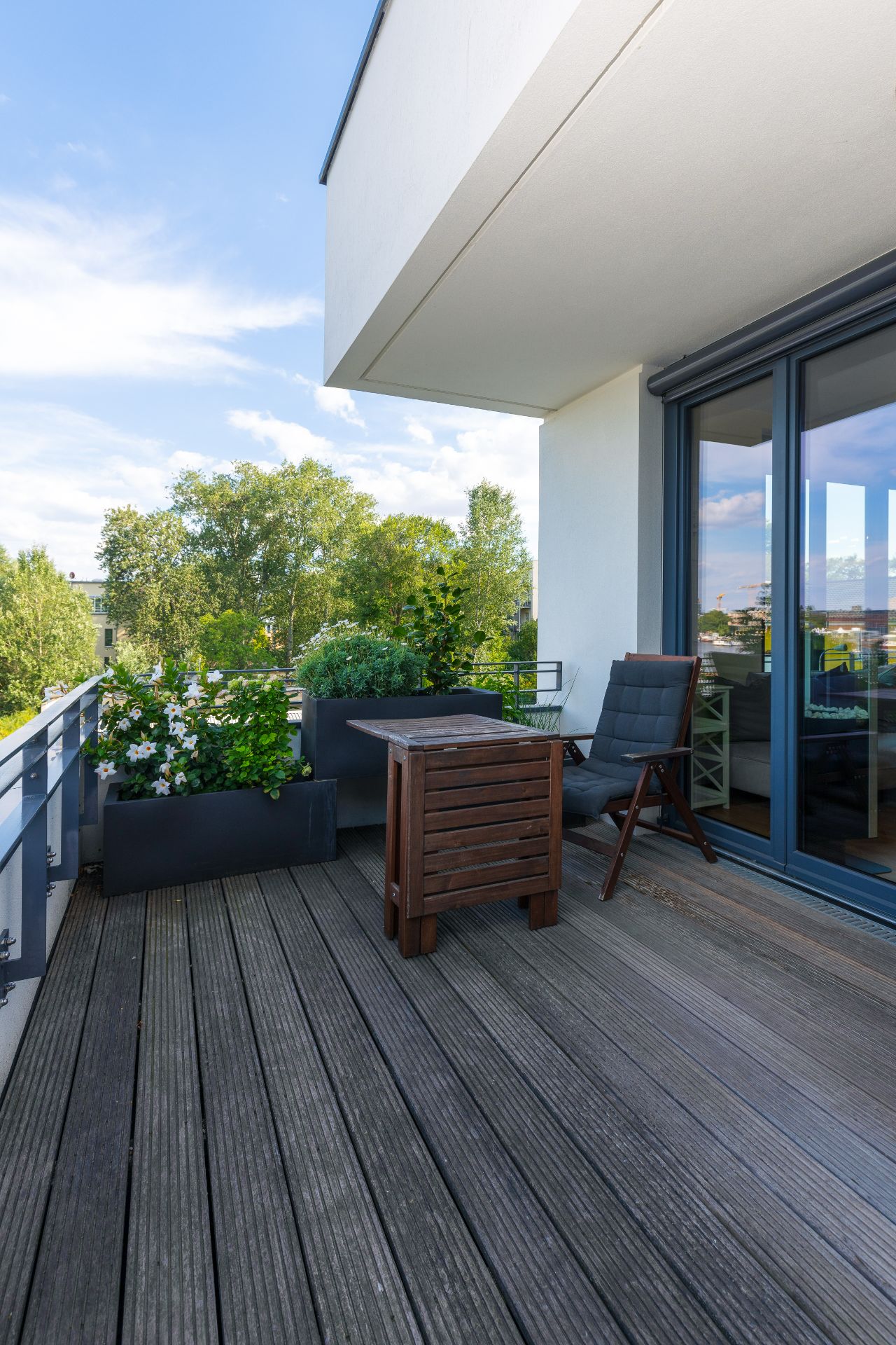 Balkon mit Holzfußboden, Pflanzkästen, kleinem Tisch und Stuhl, im Hintergrund Bäume.