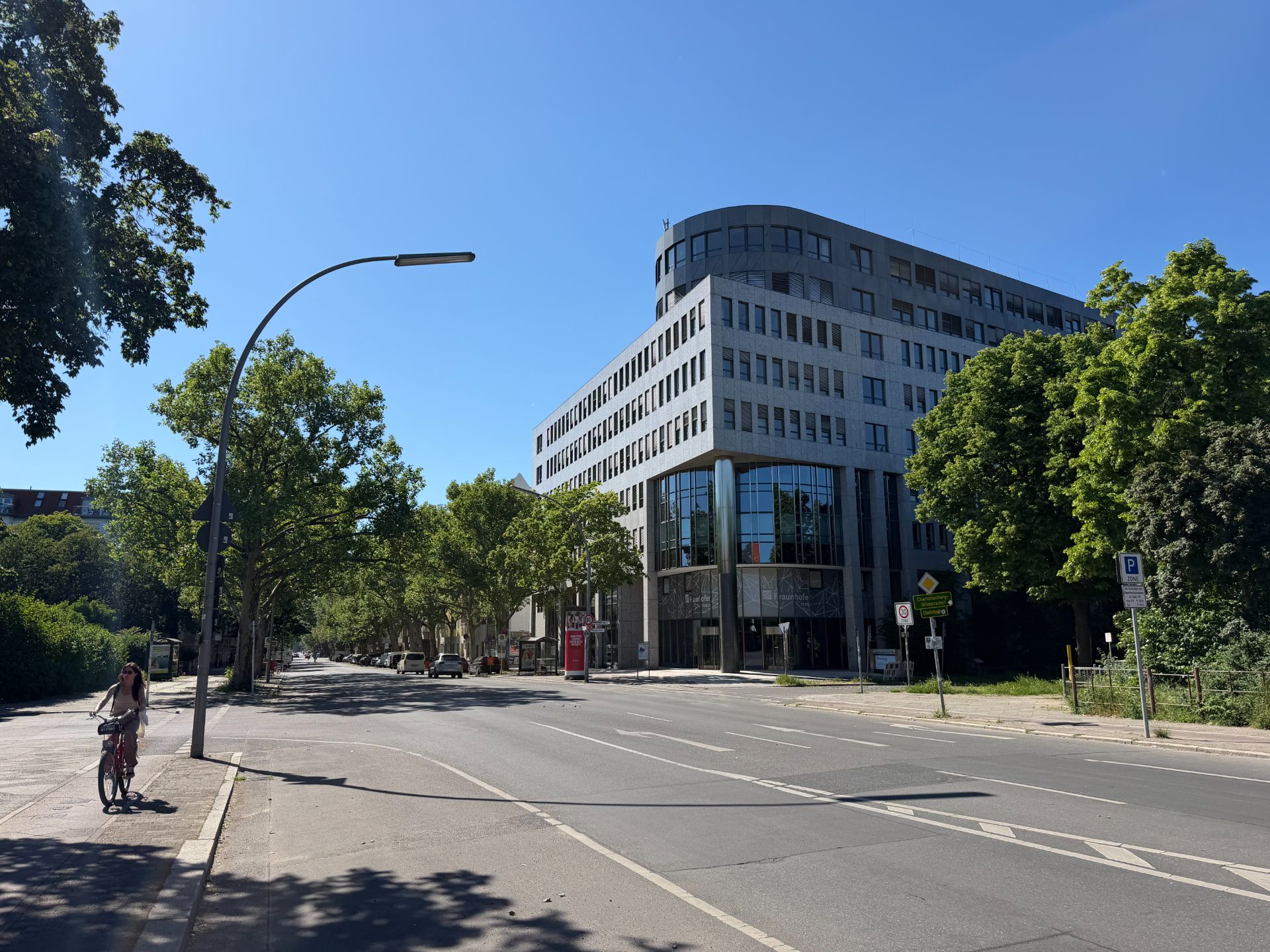 Großes modernes Gebäude an einer ruhigen Straße mit Bäumen und einer Person auf dem Fahrrad.
