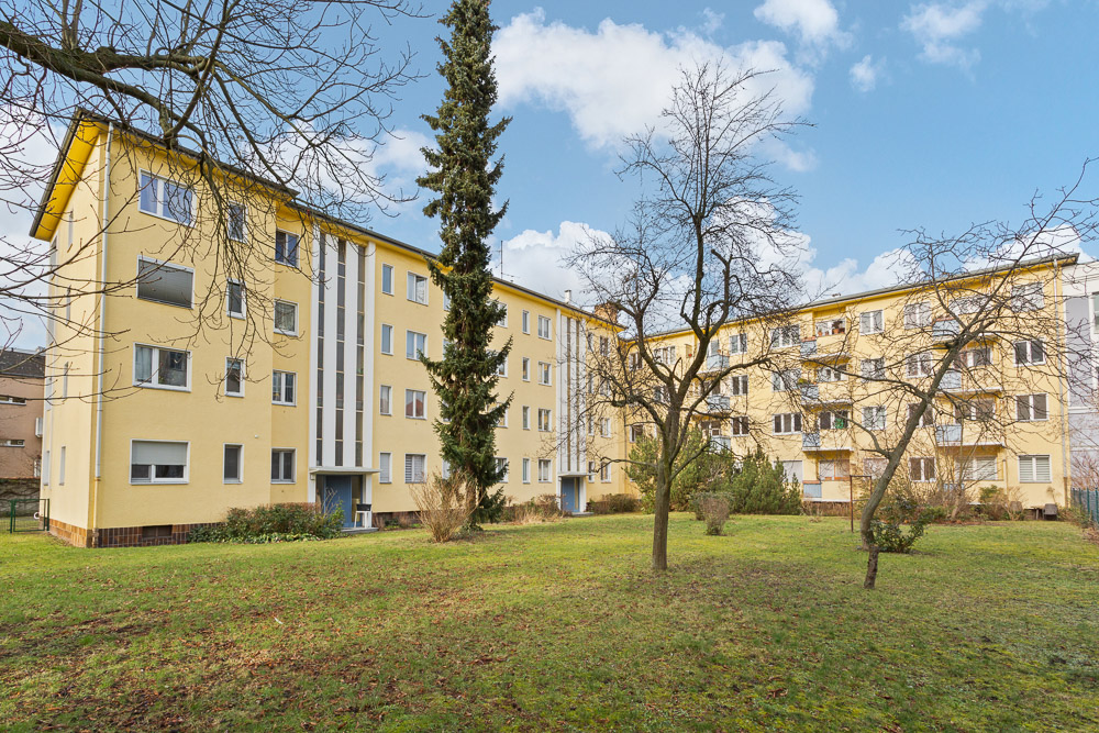 Zwei gelbe Wohngebäude mit mehreren Fenstern, davor eine Grünfläche mit Bäumen und Sträuchern.