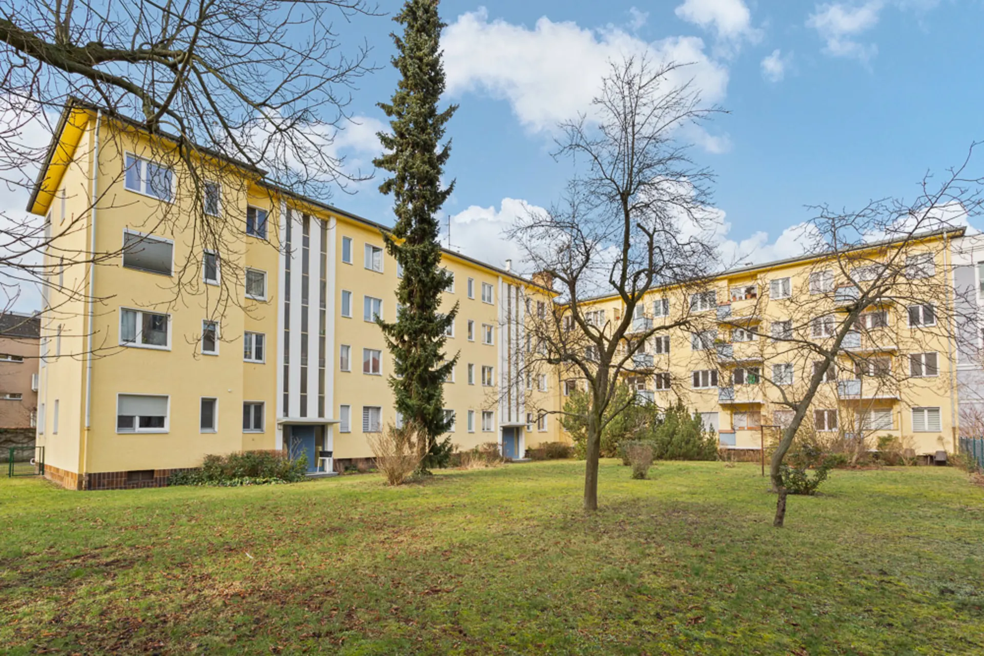 Zwei gelbe Wohngebäude mit mehreren Fenstern, davor eine Grünfläche mit Bäumen und Sträuchern.