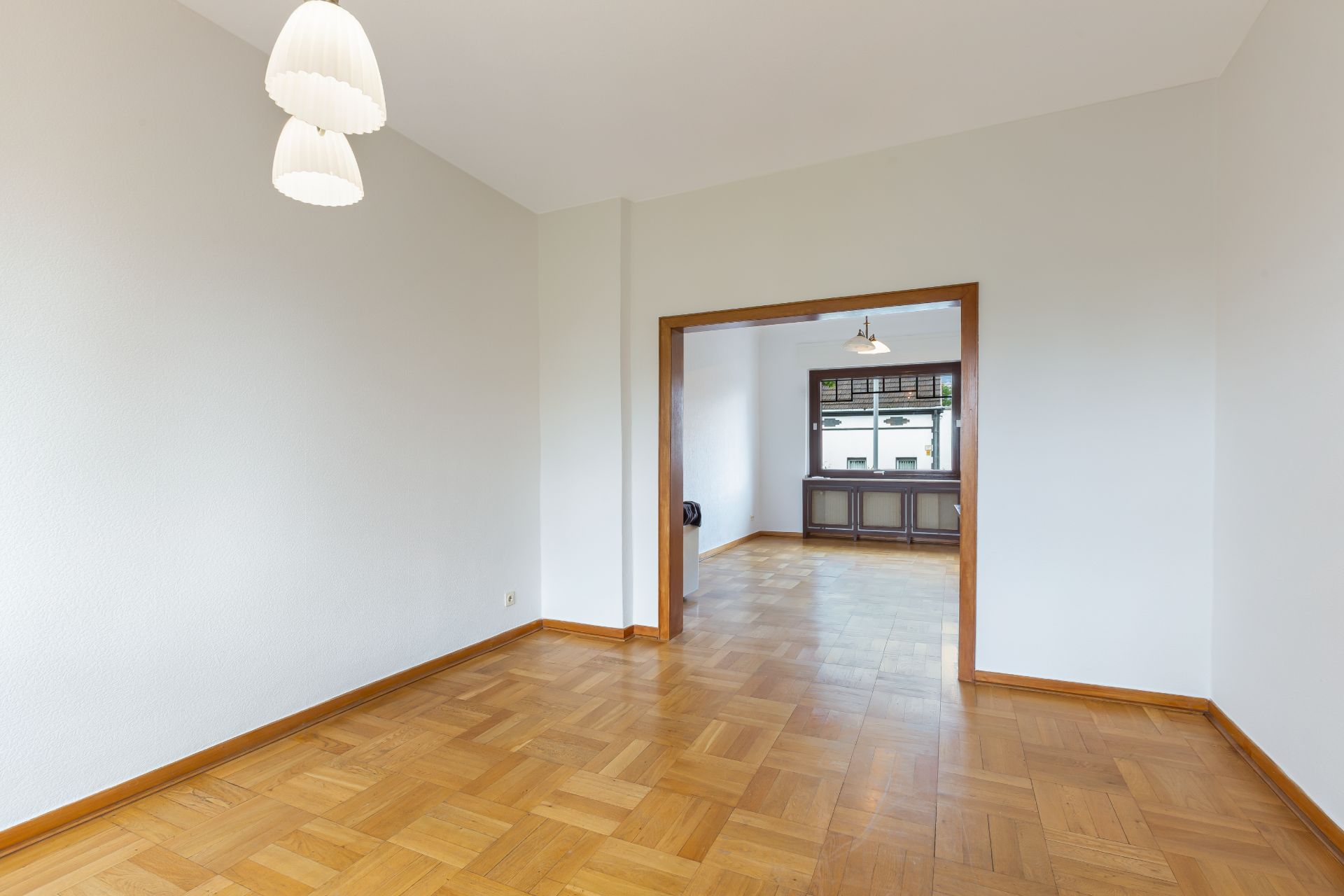 Leerer Raum mit Parkettboden, weißen Wänden und zwei Hängelampen, Blick in ein weiteres Zimmer.