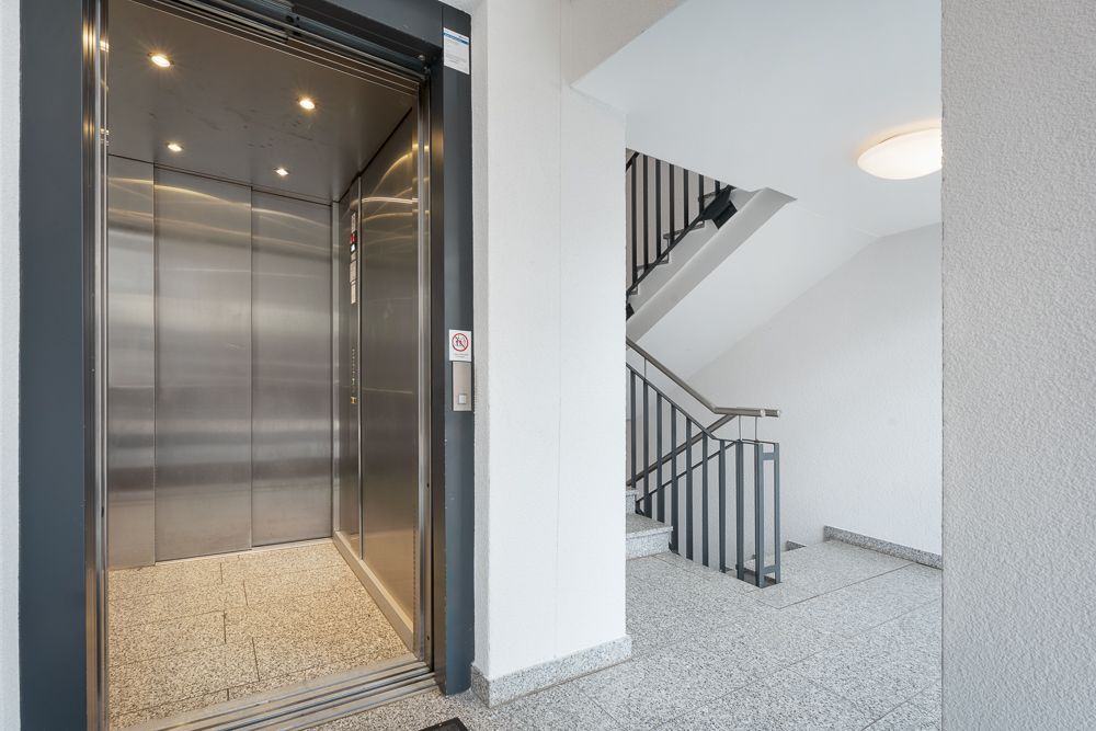 Offener Aufzug neben einem Treppenhaus in einem hellen Gebäude.