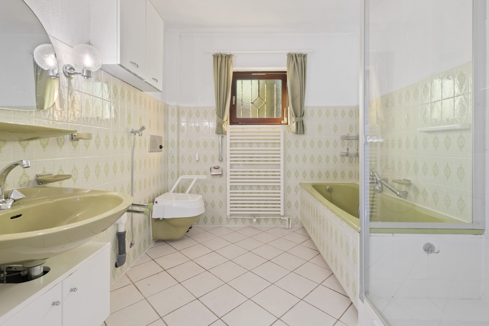 Helles Badezimmer mit grüner Badewanne, grünem Waschbecken, WC und Fenster mit Vorhängen.
