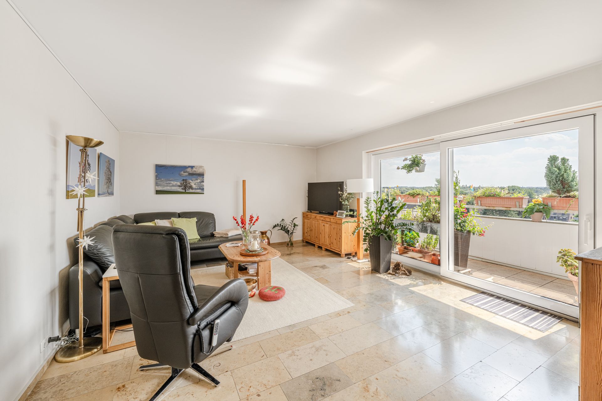 Helles Wohnzimmer mit Ecksofa, Sessel, Fernseher und Zugang zu einem Balkon mit vielen Pflanzen.