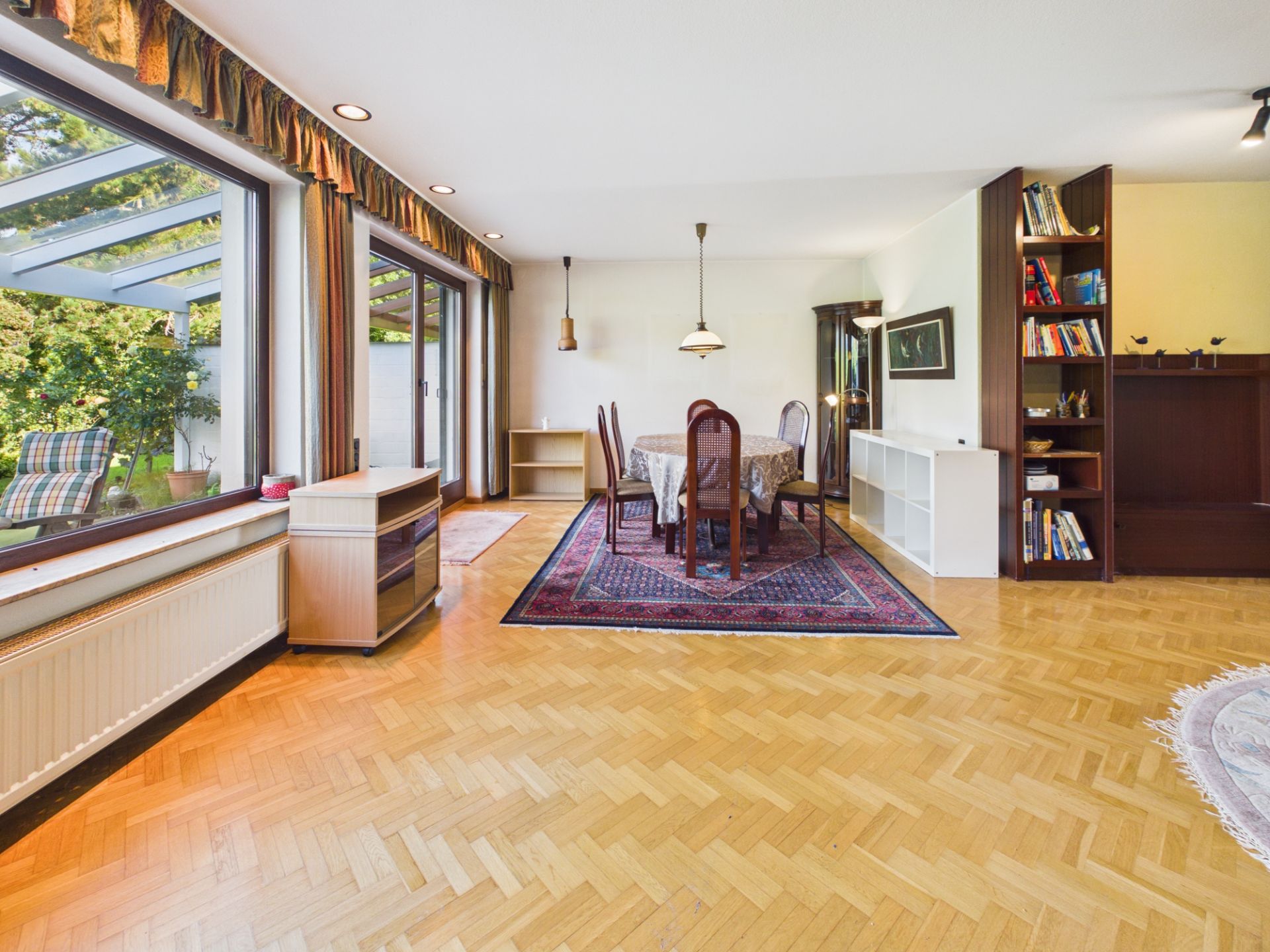 Helles Wohnzimmer mit Holzboden, großem Fenster, Esstisch, Teppich und Bücherregalen.
