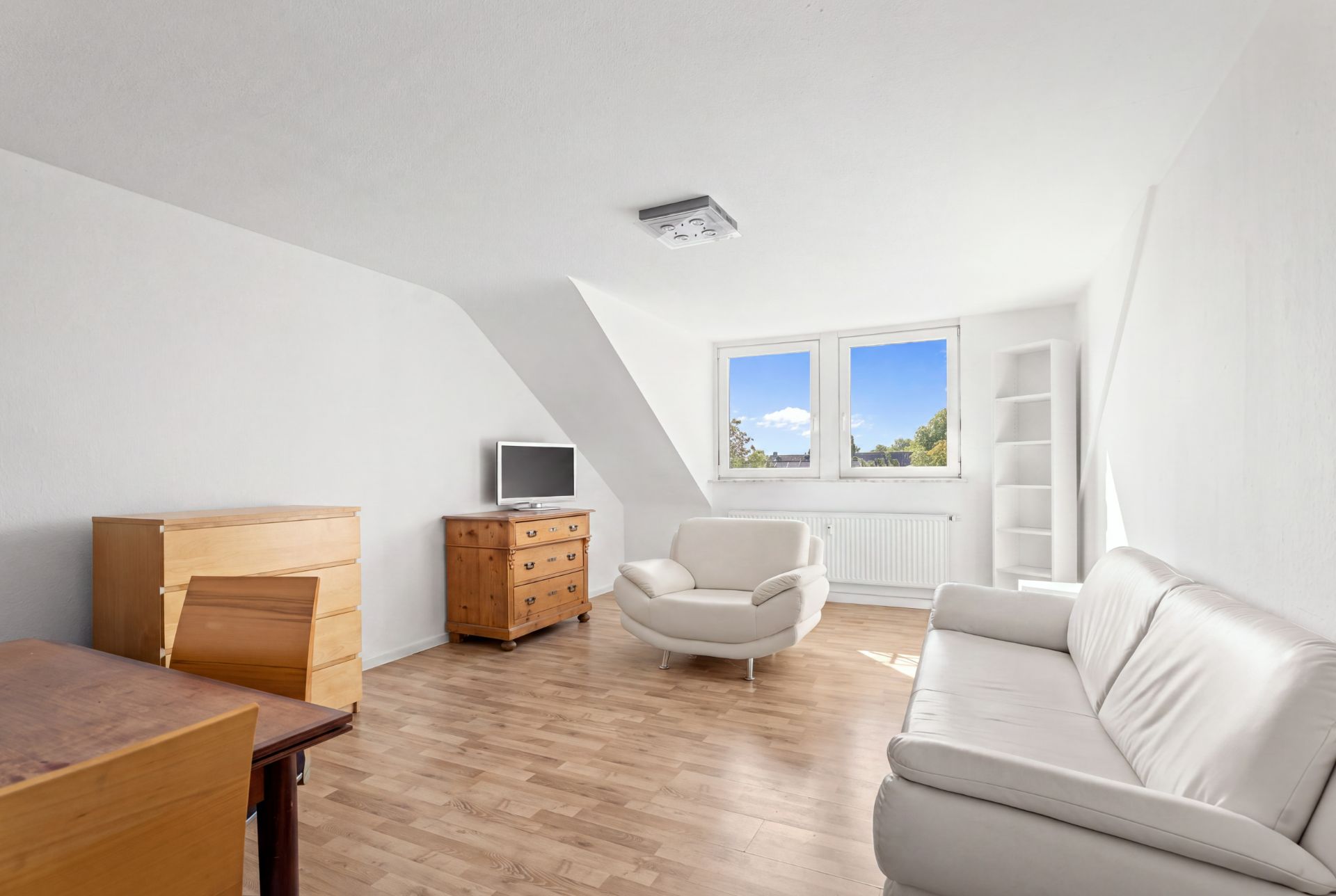 Helles Wohnzimmer mit weißem Sofa, Sessel, Holzmöbeln und zwei Fenstern.