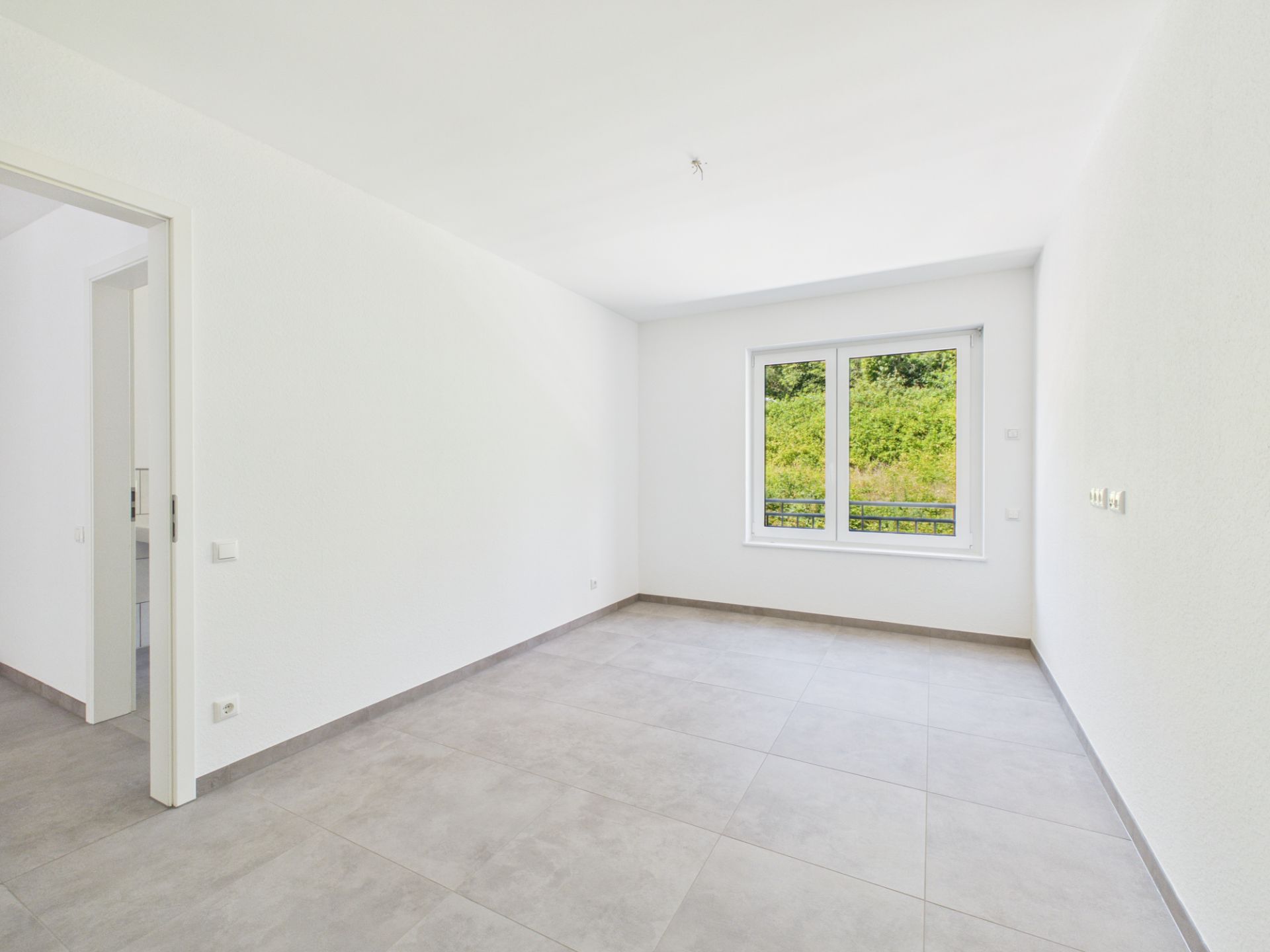 Heller leerer Raum mit großem Fenster und grauen Fliesen, Blick ins Grüne.
