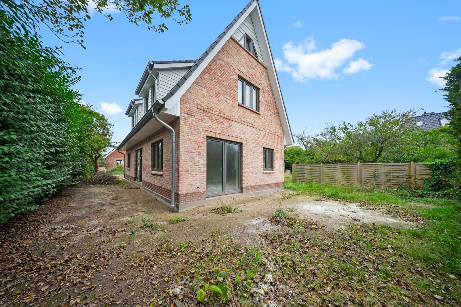 Backsteinwohnhaus mit großen Fenstern und Gartenbereich, umgeben von Bäumen und Holzzaun.