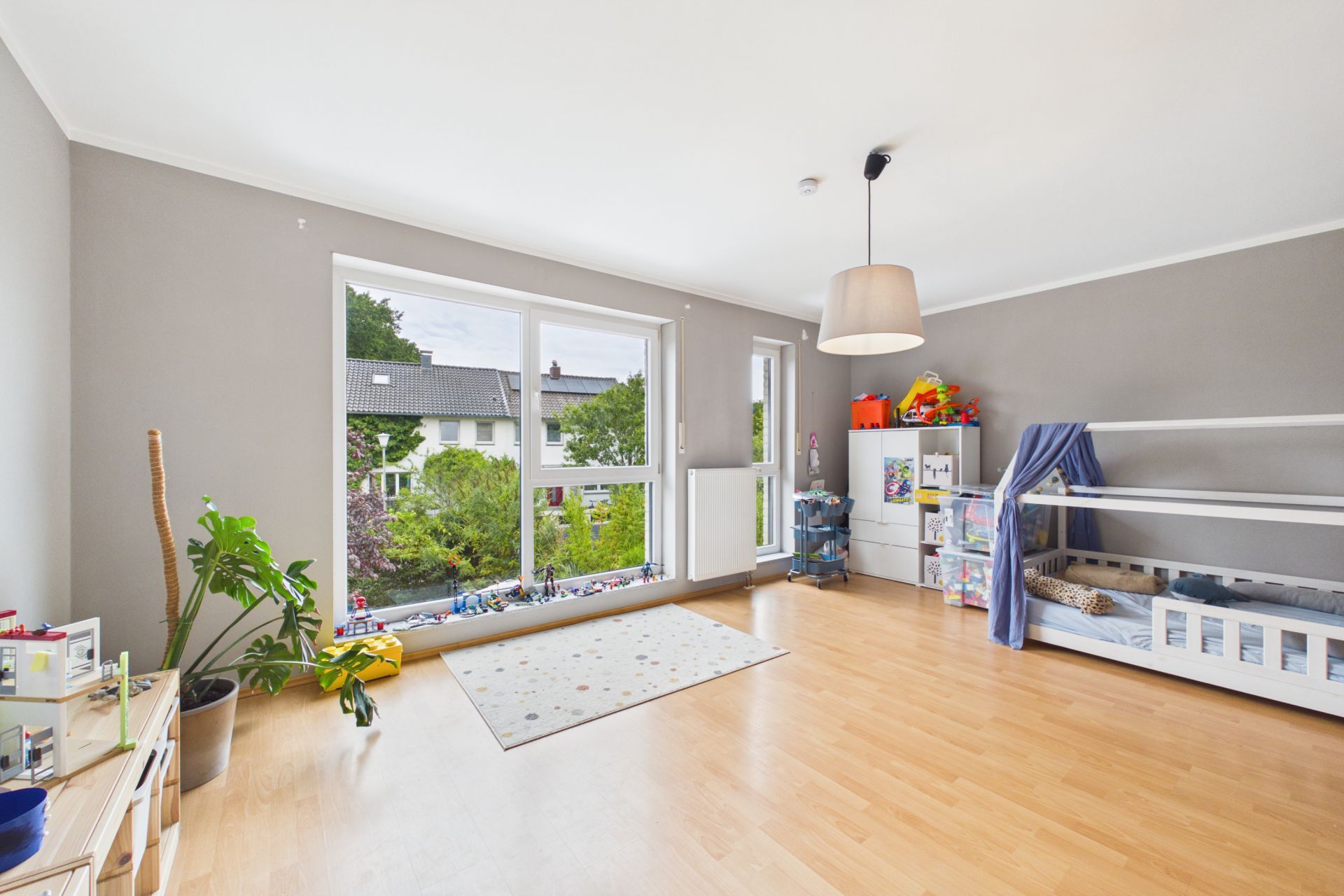 Helles Kinderzimmer mit großem Fenster, Holzboden, Spielzeug und Kinderbett.