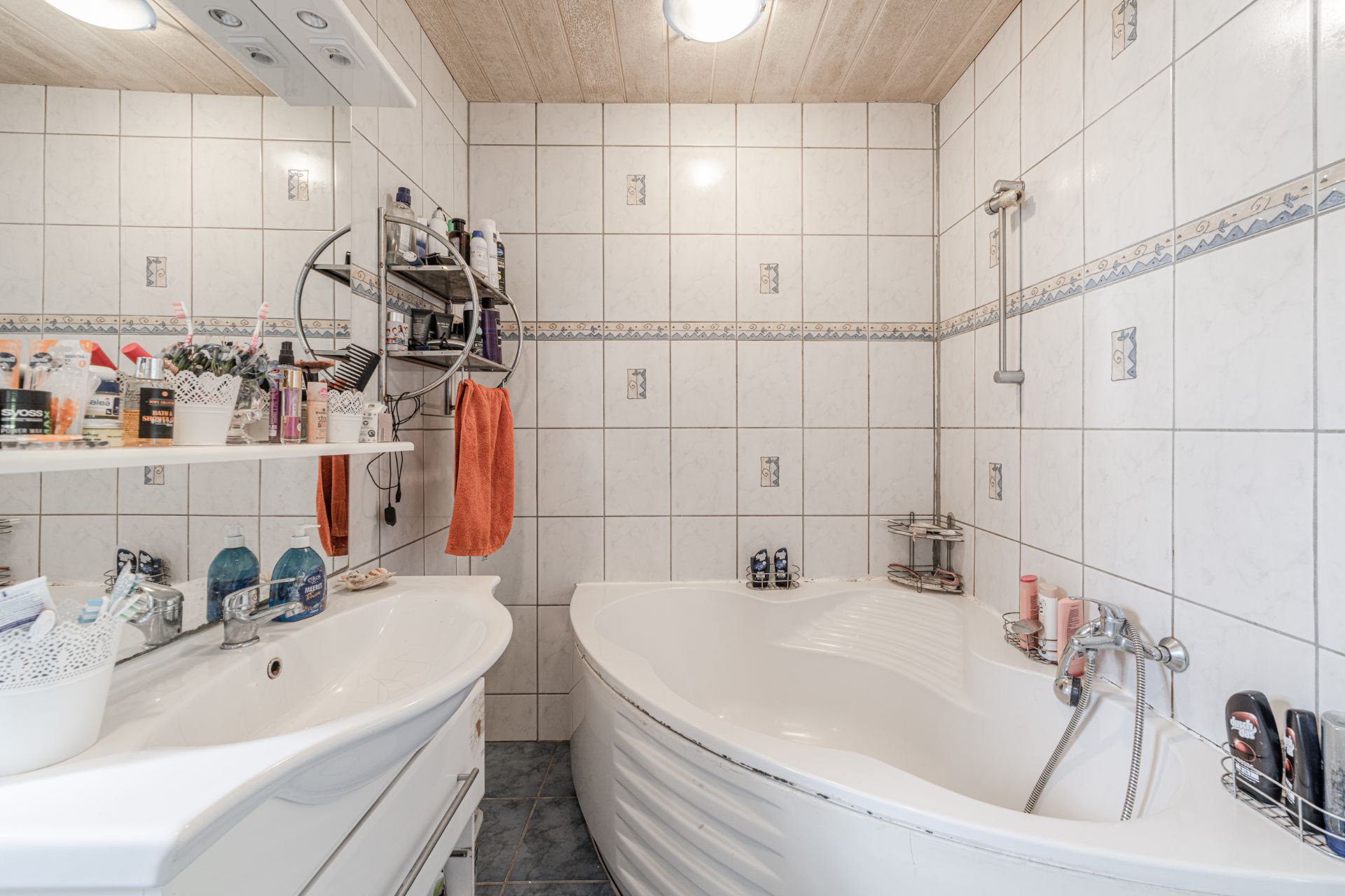 Kleines Badezimmer mit Eckbadewanne, Waschbecken, Spiegel und vielen Pflegeprodukten.