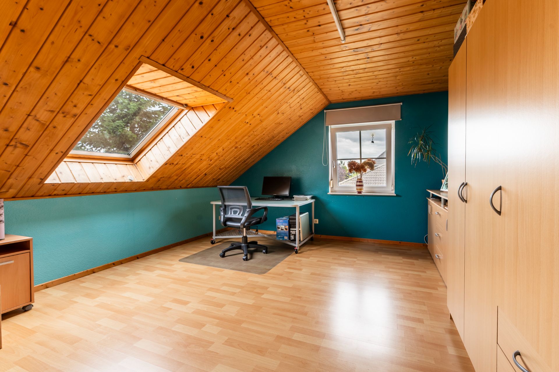 Helles Dachzimmer mit Schreibtisch, Drehstuhl, großem Schrank und Dachfenster.