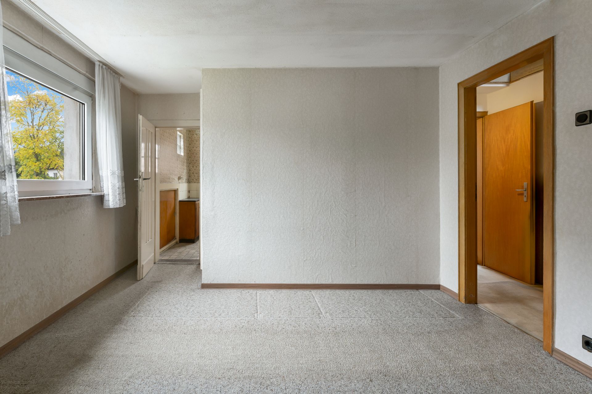 Helles Zimmer mit Teppichboden, einem Fenster, zwei offenen Türen und weißer Tapete.