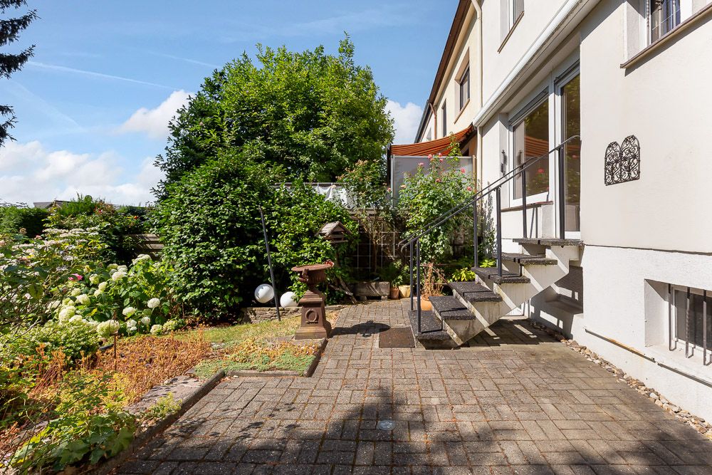 Gepflasterte Terrasse mit Treppe, umgeben von Pflanzen und Büschen neben einem Wohnhaus.