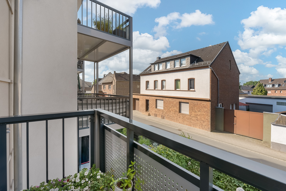 Balkon mit Blumen und Blick auf eine ruhige Wohnstraße mit Mehrfamilienhäusern.