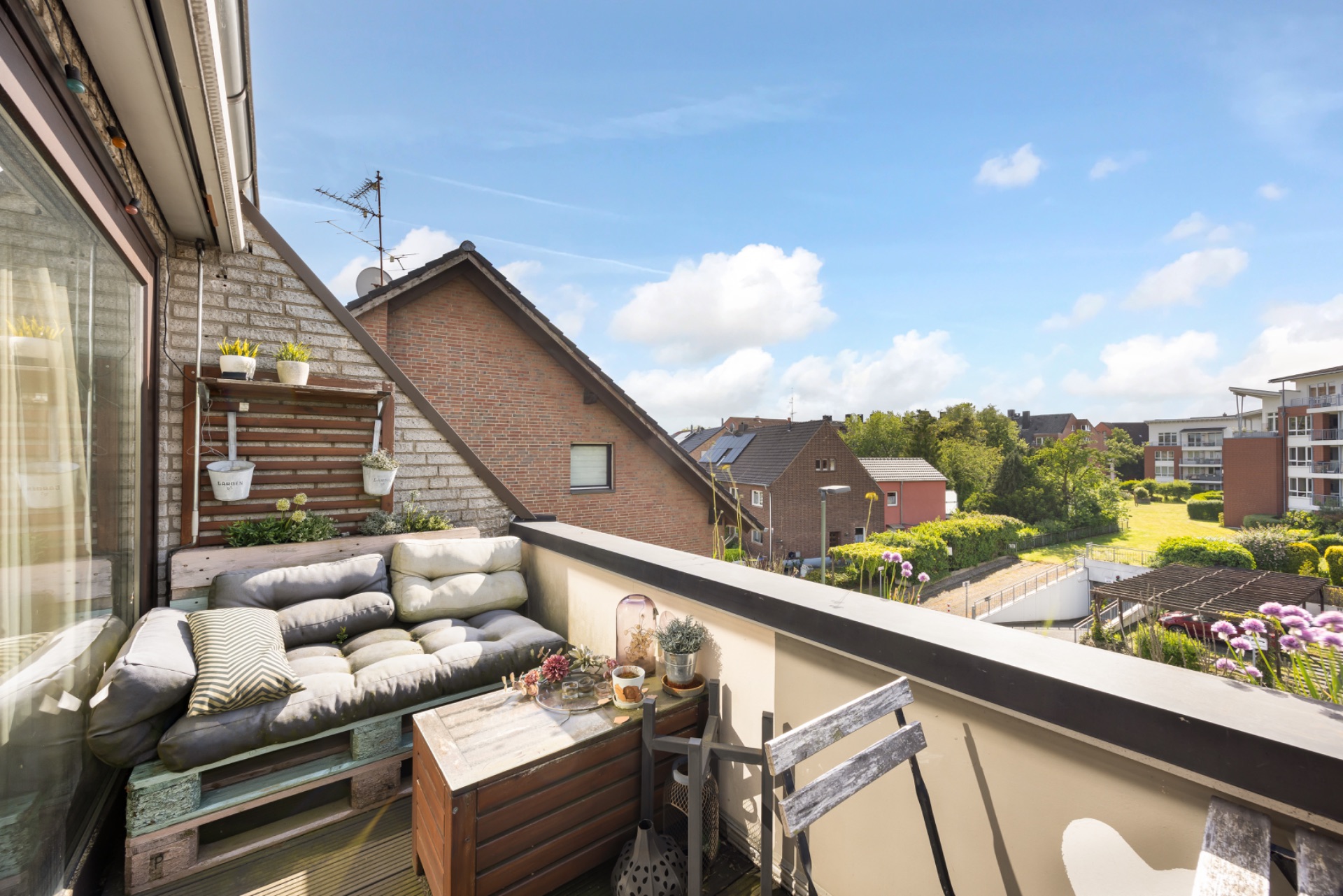 Kleiner Balkon mit Sitzbank, Holztisch, Stühlen und Pflanzen, Blick auf Nachbarhäuser und Garten.