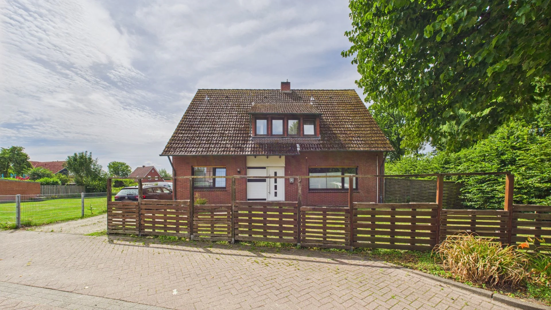 Backsteinhaus mit Satteldach und Zaun, von Bäumen und Rasen umgeben.