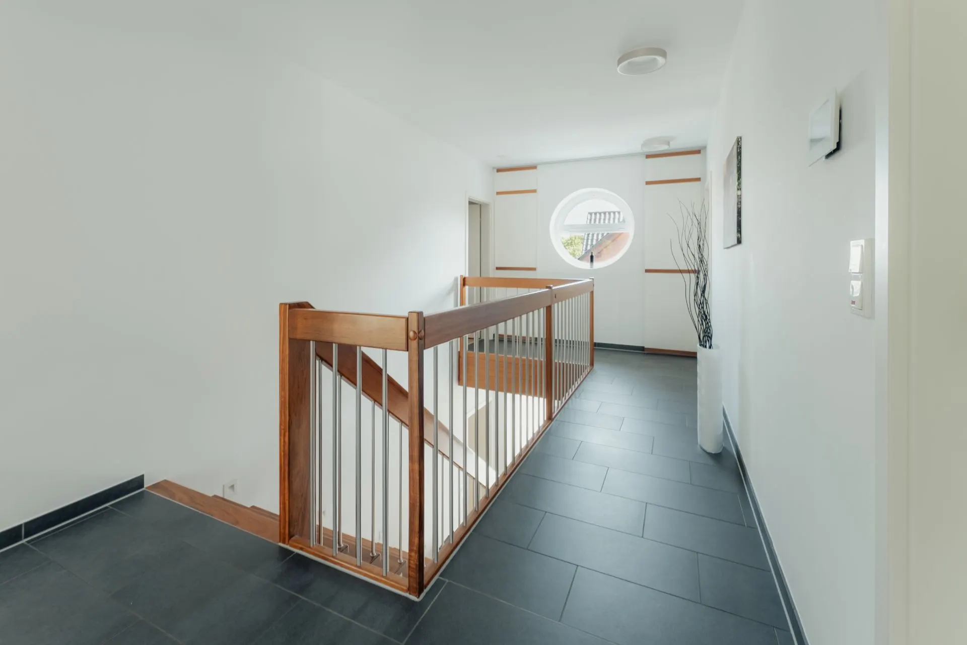 Heller Flur mit Treppengeländer aus Holz und Metall sowie rundem Fenster am Ende.