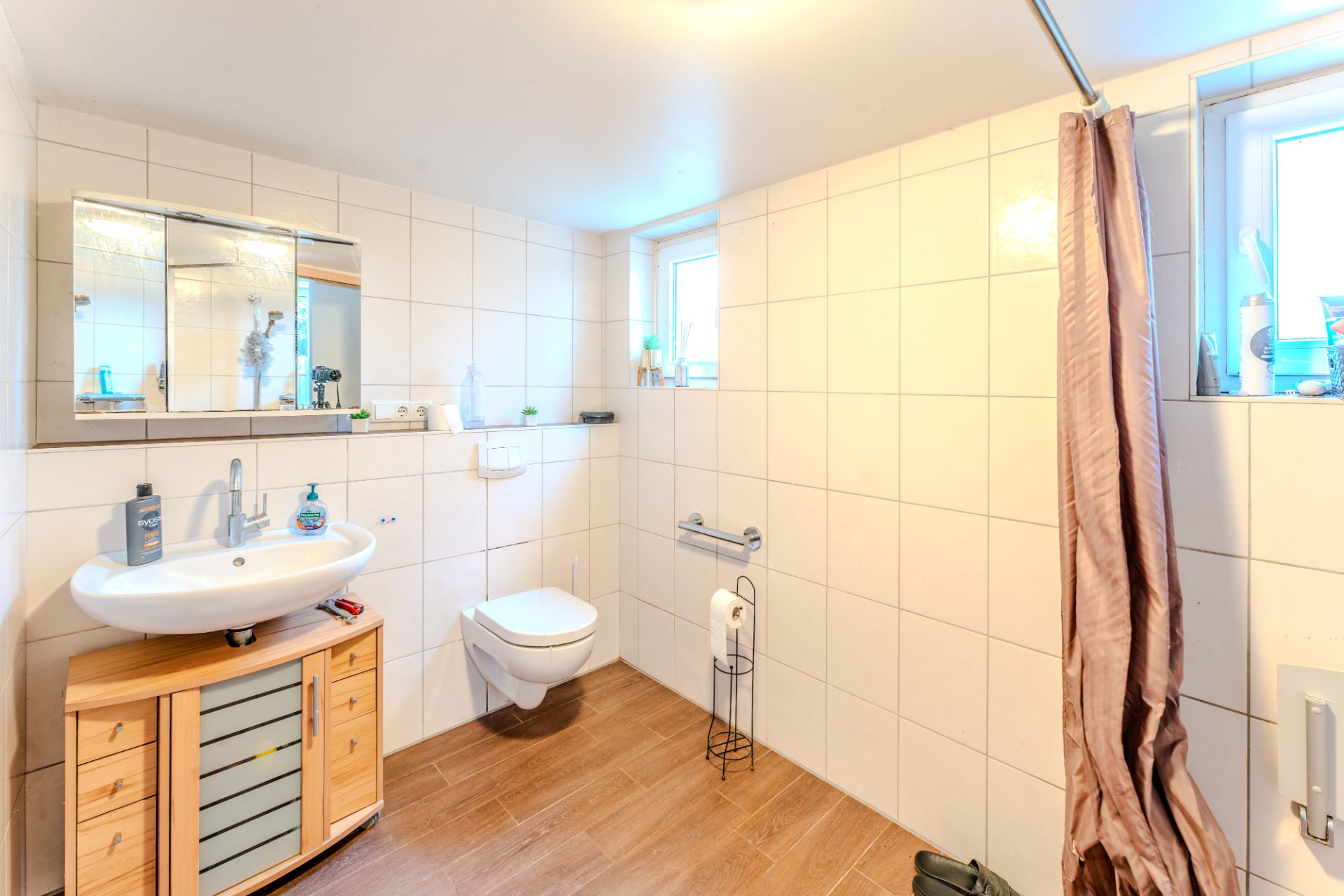 Helles Badezimmer mit Wandtoilette, Waschbecken, Spiegel und Dusche mit Vorhang.